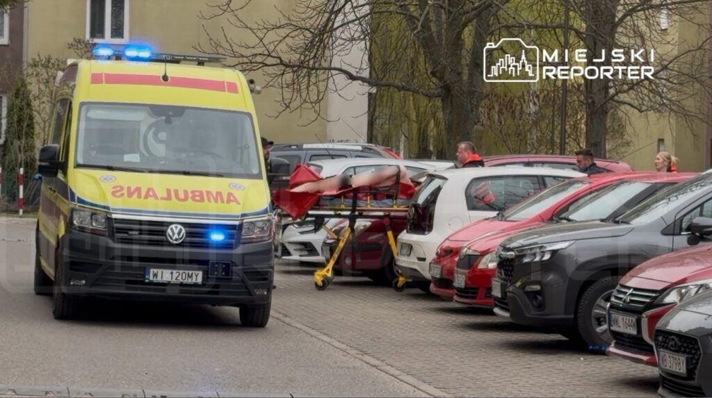 Karetka pogotowia z włączonymi sygnałami świetlnymi znajduje się na parkingu, obok leżącego na noszach poszkodowanego.