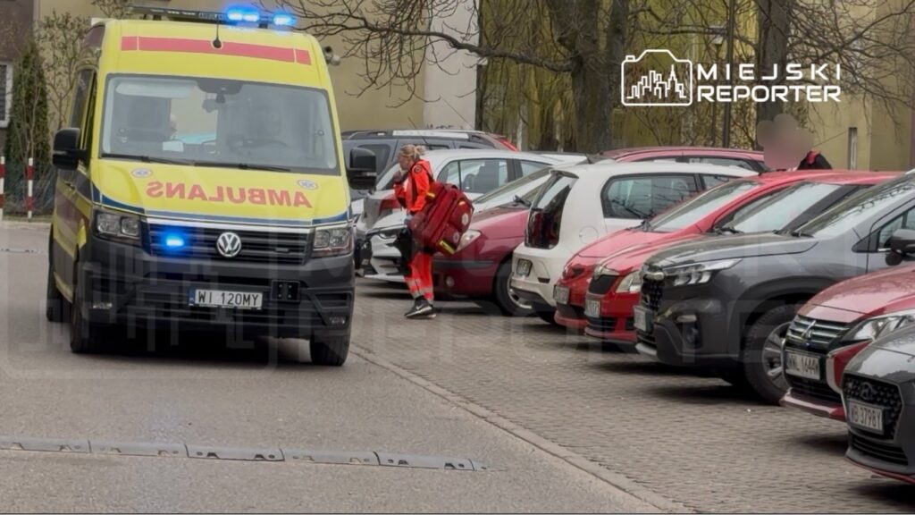 Ratownik medyczny w czerwonym stroju niesie torbę do karetki na parkingu, obok zaparkowanych samochodów.