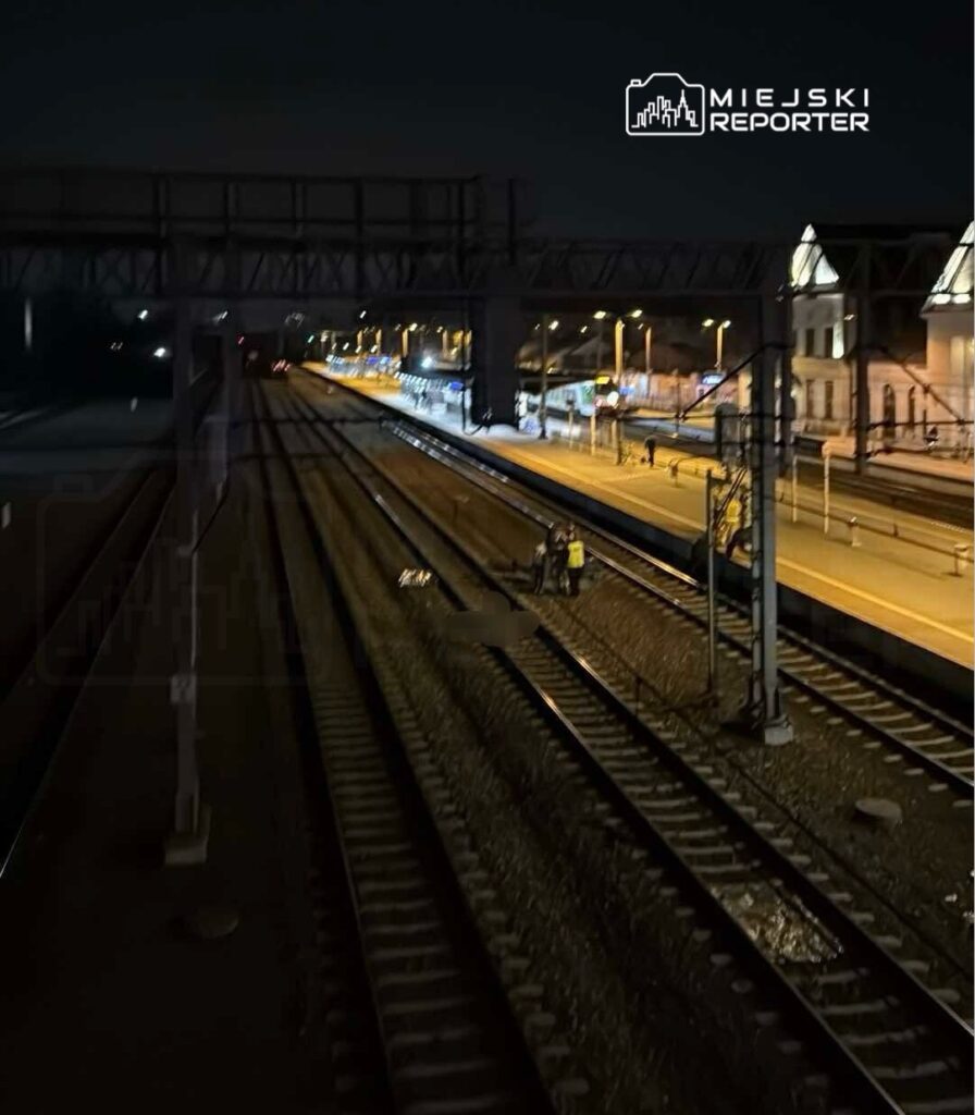 Grupa osób w odblaskowych kamizelkach pracuje na torach kolejowych w nocy, przy oświetlonej stacji.