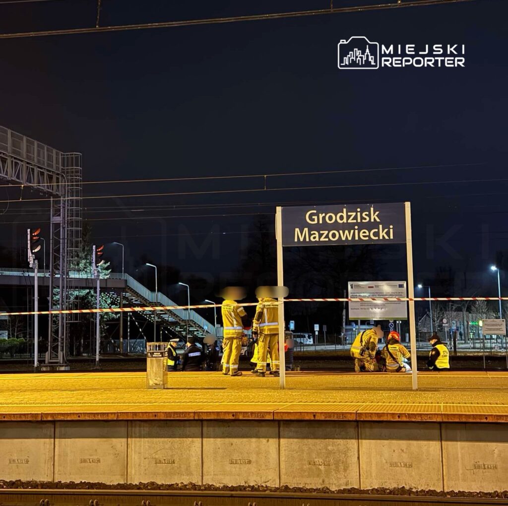 Grupa pracowników w odblaskowych kamizelkach naprawia torowisko na stacji Grodzisk Mazowiecki w nocy.