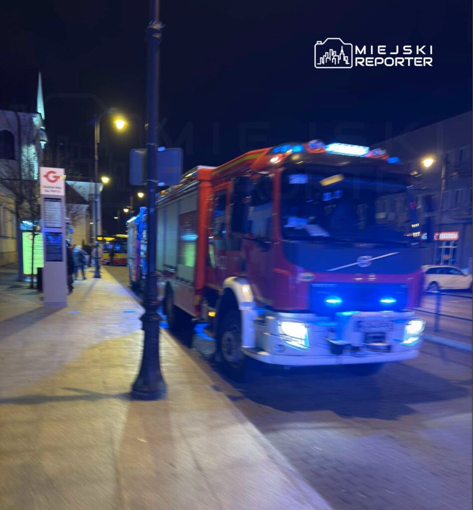 Czerwony wóz strażacki z niebieskimi światłami zaparkowany na ulicy w nocy, obok przechodniów.