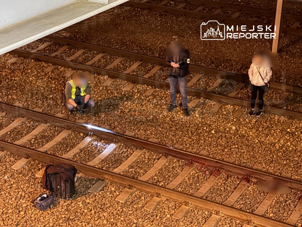 Mężczyzna w kamizelce odblaskowej sprawdza tory kolejowe, a dwie osoby stoją obok na podłożu z kamieni.