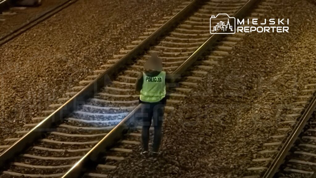 Policjant w odblaskowej kamizelce sprawdzający tory kolejowe w nocy, otoczony żwirkiem i torami.