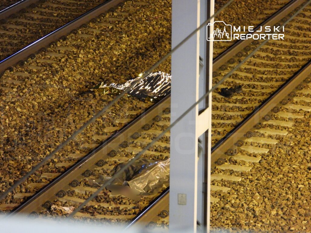 Obiekt w folii leży na torach kolejowych, otoczony kamieniami. W tle widoczna jest infrastruktura kolejowa.