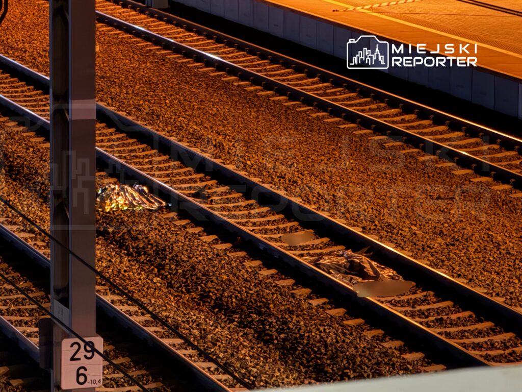 Na torach kolejowych leży folia termiczna w pobliżu przystanku, otoczona kamieniami.