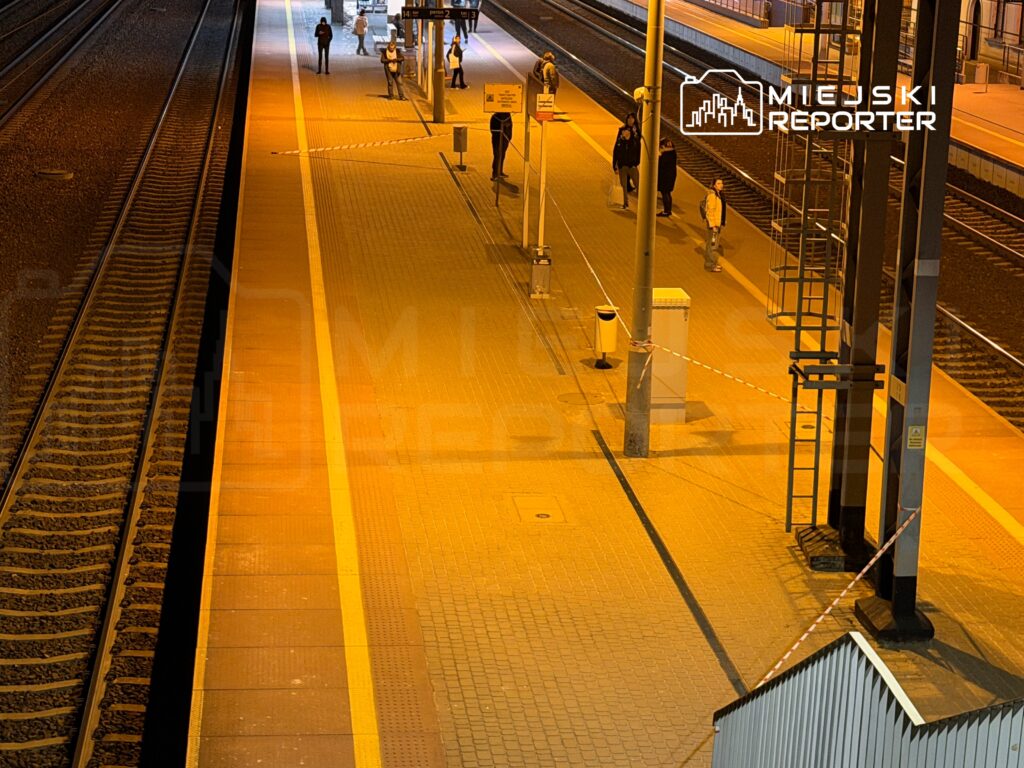 Pasażerowie czekają na peronie oświetlonym pomarańczowym światłem, obok torów kolejowych.