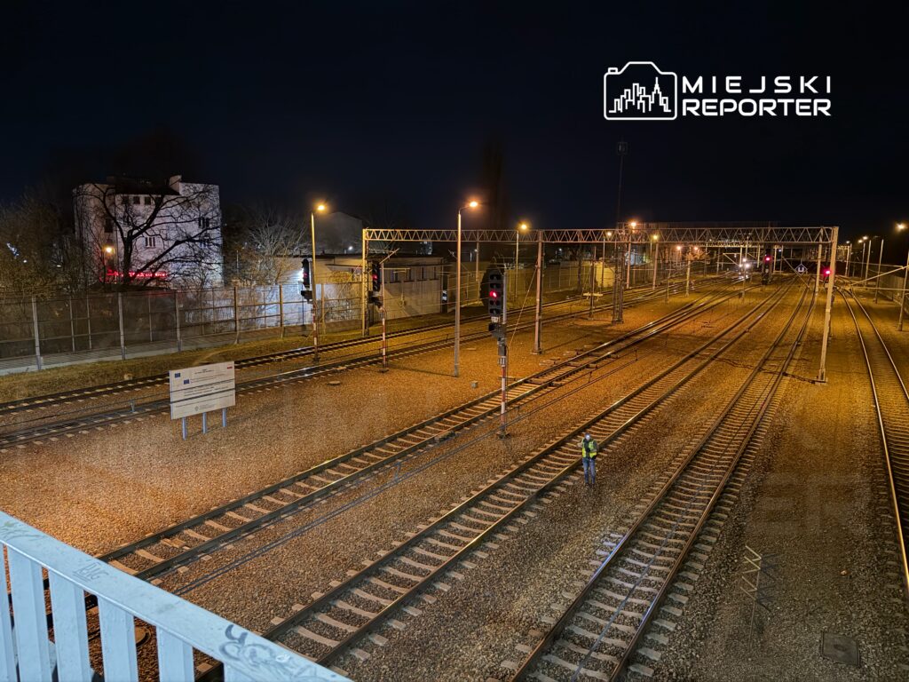 Pracownik w kamizelce odblaskowej sprawdza tory kolejowe w nocnym otoczeniu, przy oświetleniu lamp.