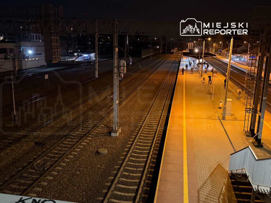 Grupa ludzi spacerująca po peronie stacji kolejowej w nocy, otoczenie oświetlone lampami.