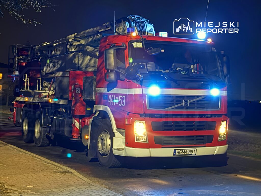 Czerwony wóz strażacki z podnośnikiem zaparkowany na ulicy w nocy, z włączonymi sygnałami świetlnymi.