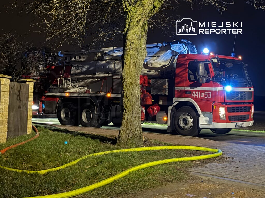 Czerwony wóz strażacki z podnośnikiem parkowany przy drodze, z wężami gaśniczymi rozłożonymi na trawie.