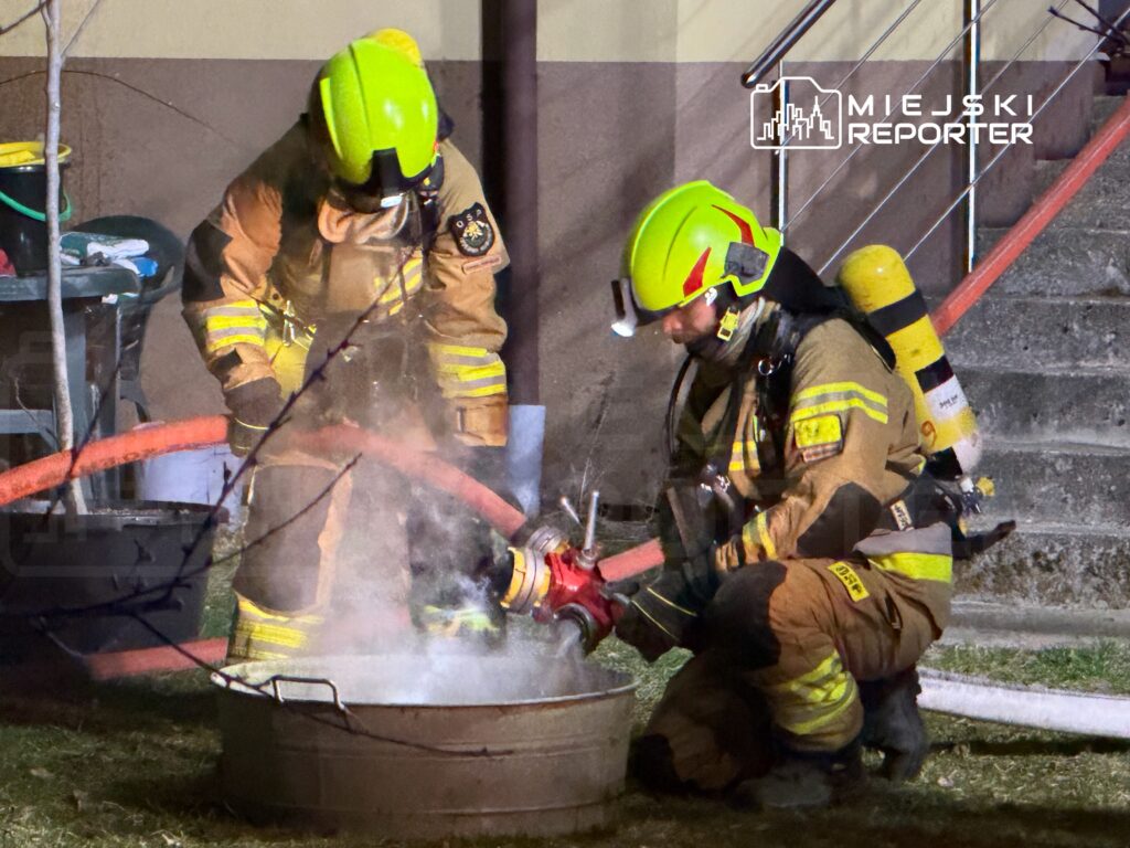Dwaj strażacy w hełmach i mundurach gaszą ogień w metalowej misce, używając węża strażackiego.