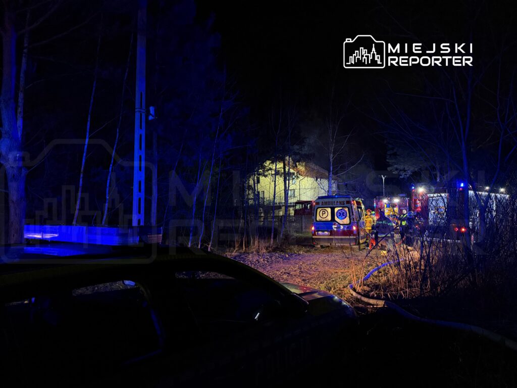 Ratownicy w mundurach przygotowują sprzęt w lesie, obok pojazdów straży pożarnej i karetki, w nocy.