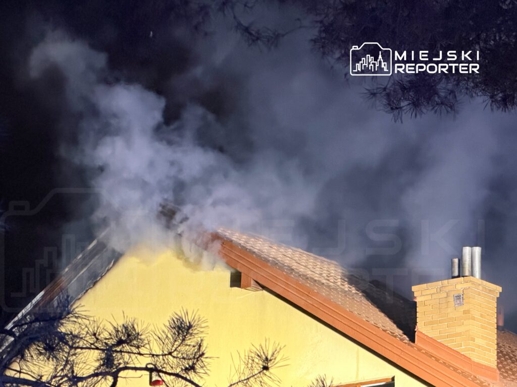Z dachu żółtego budynku wydobywa się gęsty dym, w tle ciemne niebo i drzewa.