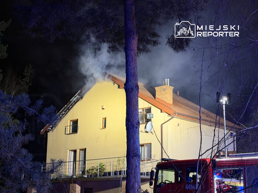Dym wydobywający się z dachu żółtego domu jednorodzinnego w nocy, obok zaparkowany wóz strażacki.