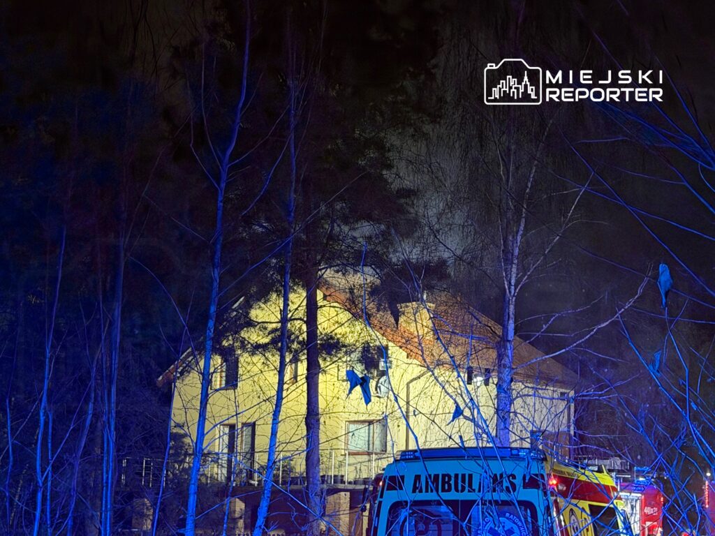 W nocy, w lesie, przy oświetlonym budynku widać karetki pogotowia oraz pojazdy straży pożarnej.
