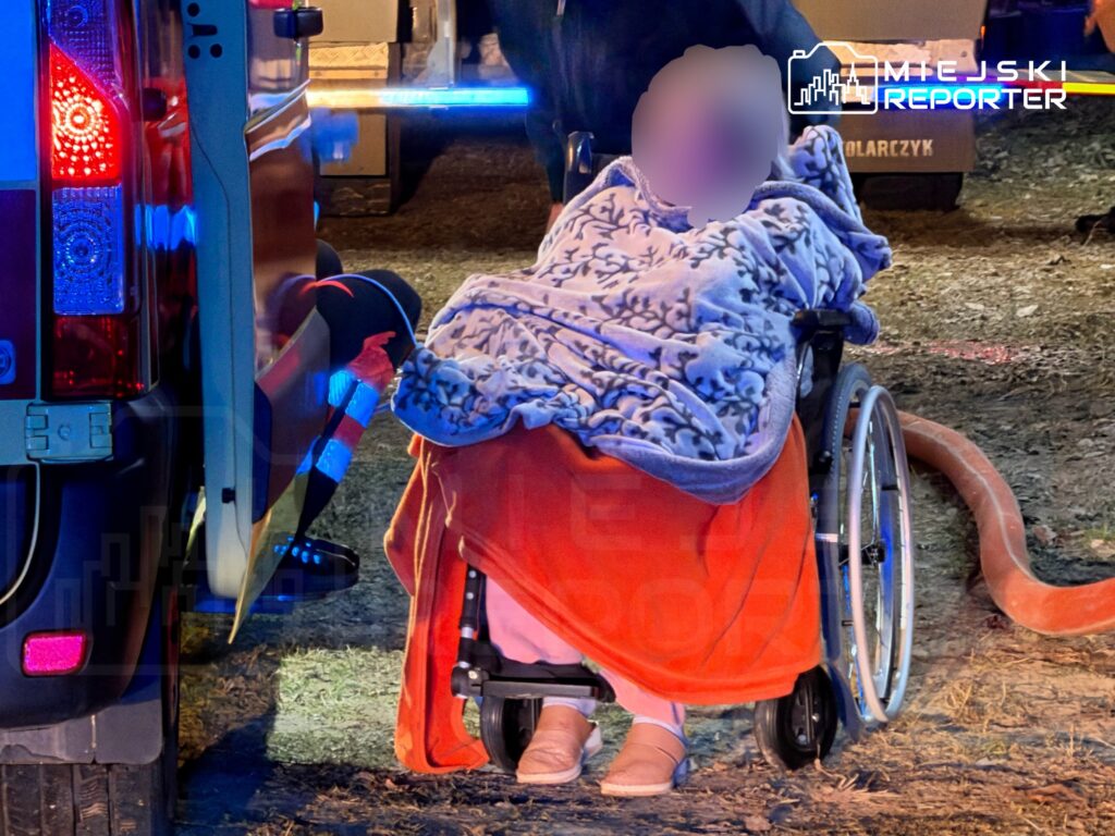 Kobieta w wózku inwalidzkim owinięta kocem, czeka na pomoc medyczną przy ambulansie w ciemnym otoczeniu.