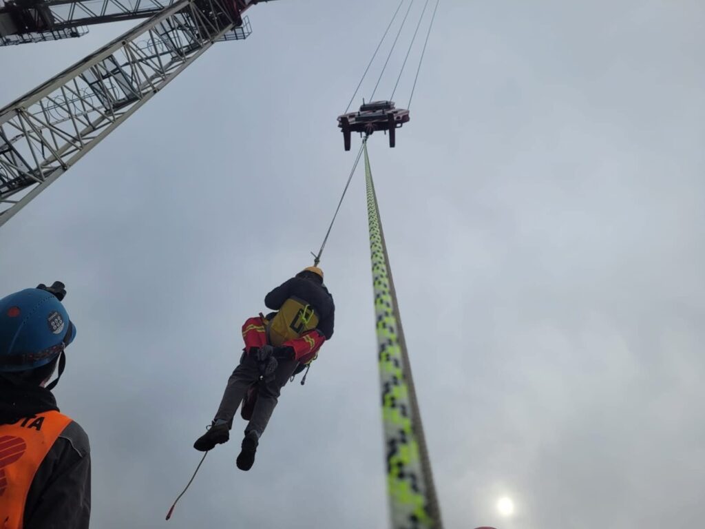 Ratownik w sprzęcie ochronnym opuszczający się na linie z dźwigu w pochmurnym otoczeniu.