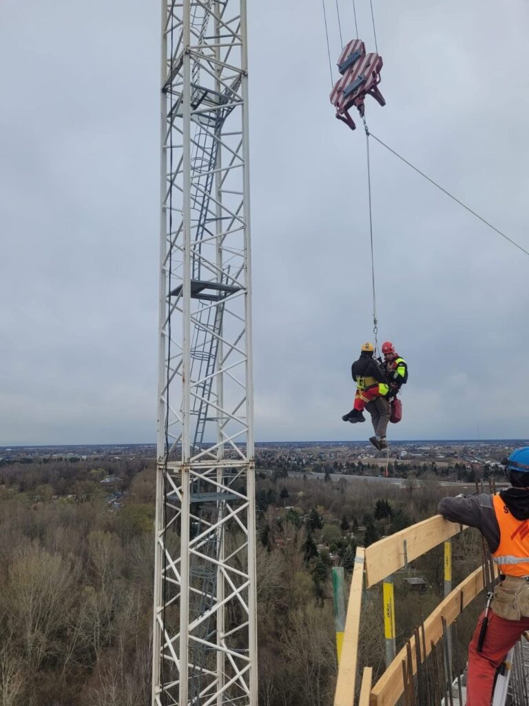 Robotnicy w kaskach podwieszają osobę na linach obok wieży budowlanej, z widokiem na okoliczne tereny.