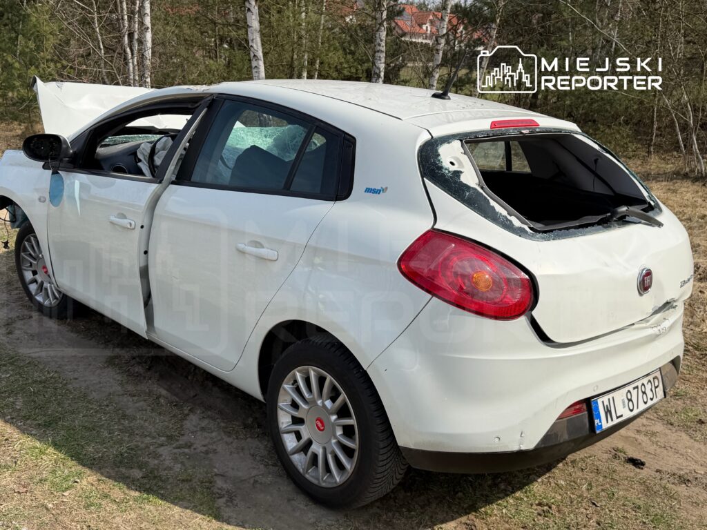 Uszkodzony biały samochód osobowy z otwartymi drzwiami stoi na trawie w lesie, z wybitym tylnym szybą.