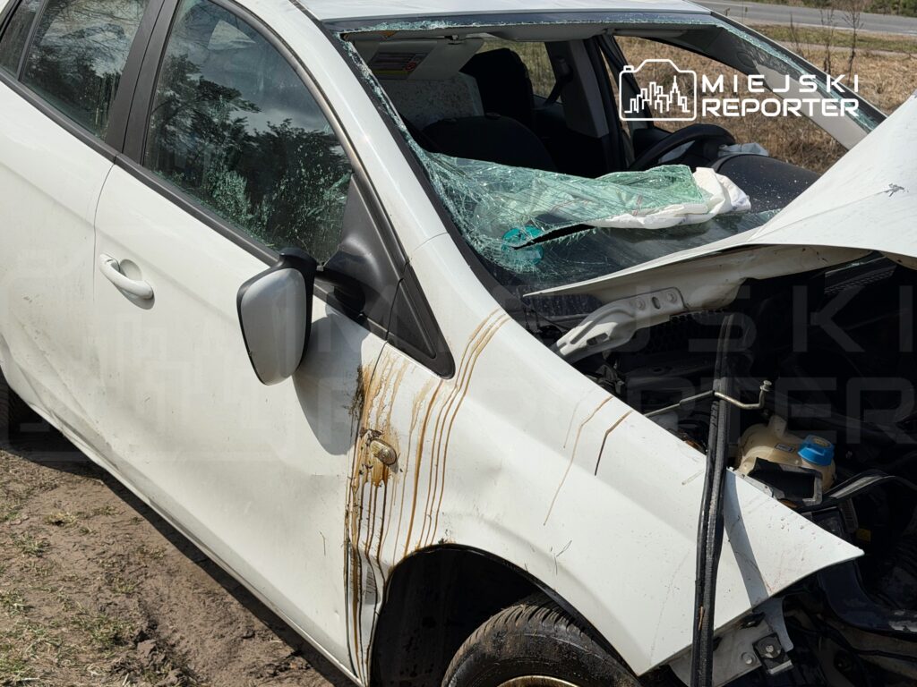 Uszkodzony biały samochód osobowy zaparkowany na poboczu drogi, z rozbitą szybą i widocznymi śladami błota.