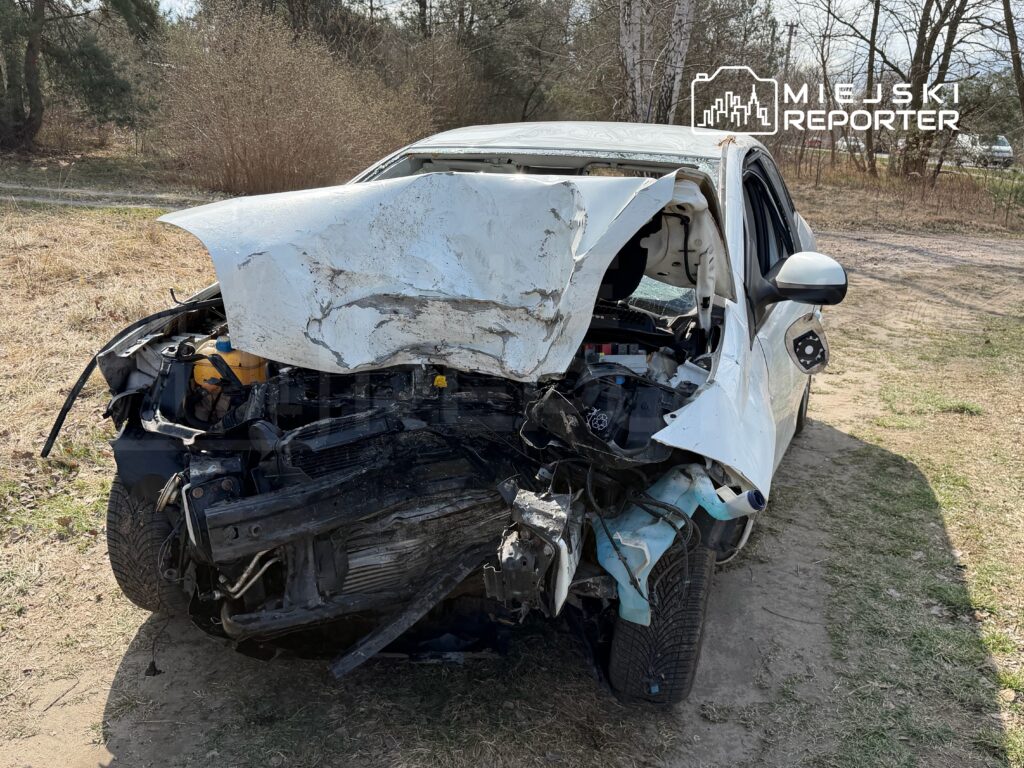 Zniszczony samochód osobowy z wgniecionym przodem stoi na trawie w otoczeniu drzew.