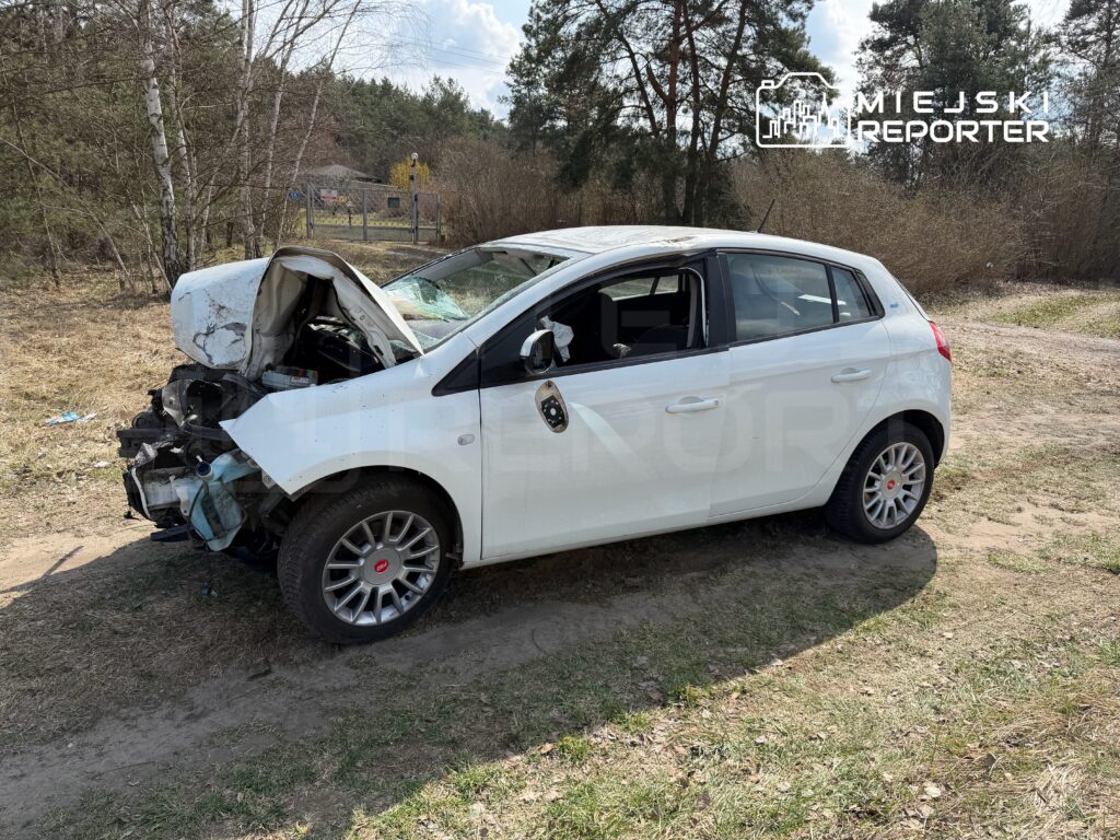 Uszkodzony biały samochód osobowy stoi na poboczu drogi, z otwartą maską i widocznymi zniszczeniami przodu. W tle drzewa i ogrodzenie.