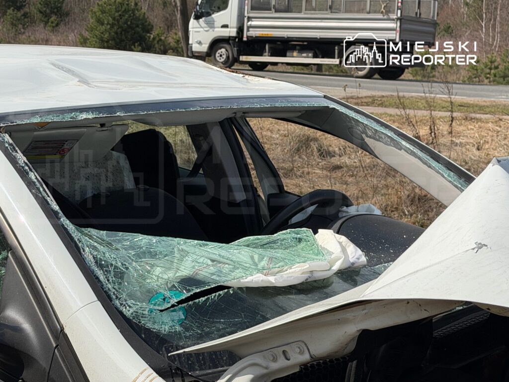 Połamana szyba samochodowa z rozbitym dachem, stojąca na poboczu drogi w otoczeniu drzew.