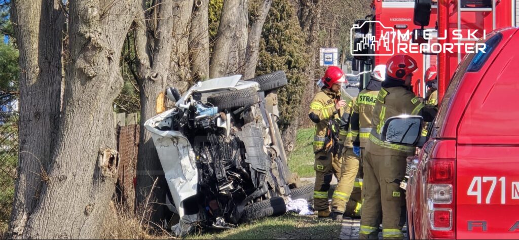 Wozidło przewrócone na bok obok drzew, a strażacy w mundurach omawiają sytuację na miejscu wypadku.