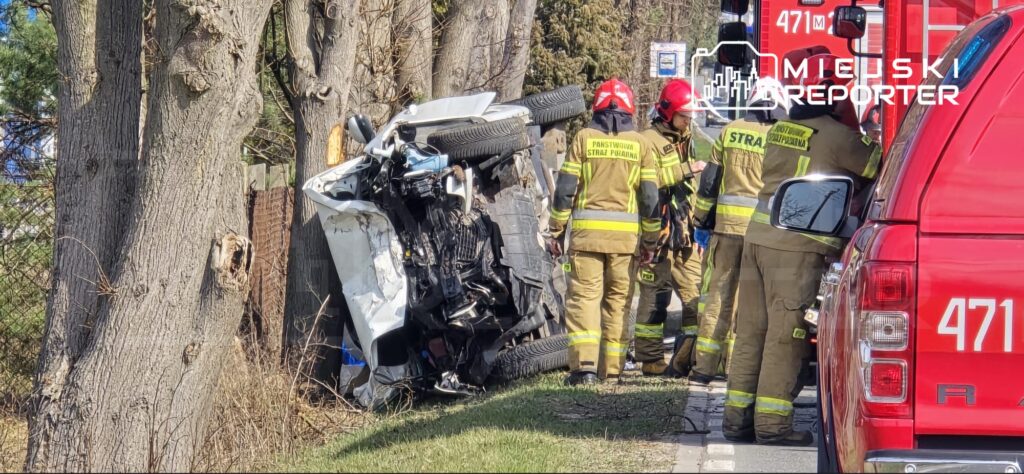 Grupa strażaków w mundurach pomaga przy przewróconym pojeździe leżącym na boku obok drogi.