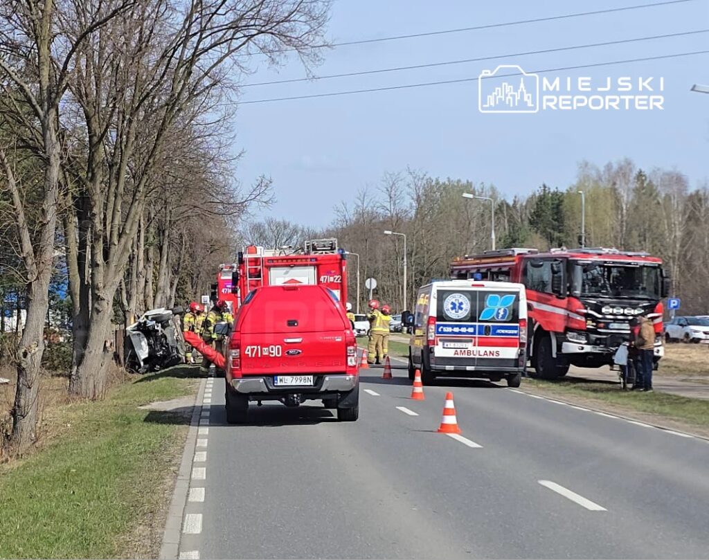 Pojazd w rowie, obok strażacy i karetka, na drodze z ustawionymi pachołkami ostrzegawczymi.