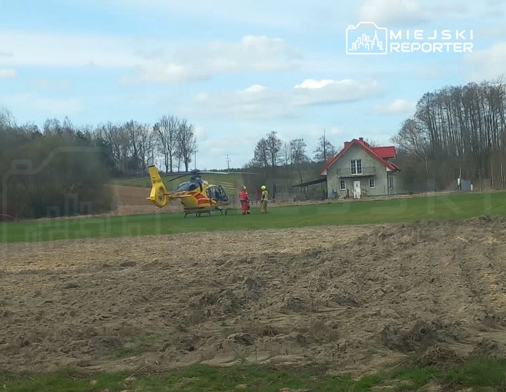 Śmigłowiec ratunkowy ląduje na zielonym terenie obok domu, a ratownicy przygotowują się do akcji.