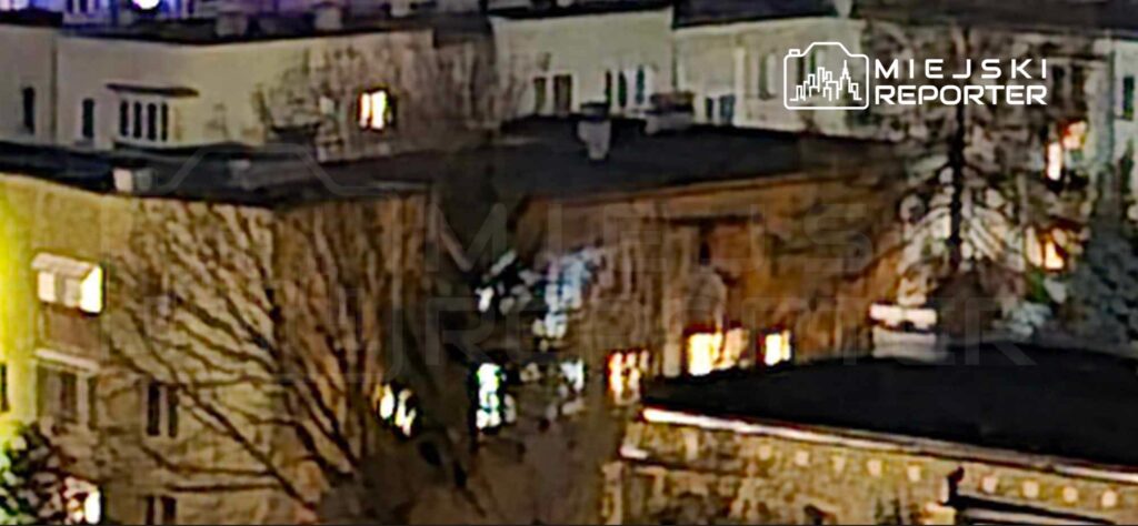 Oświetlony budynek mieszkalny z widocznymi oknami w nocy, otoczony nagimi drzewami.