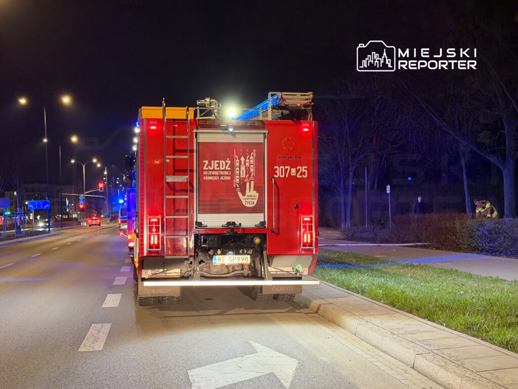 Czerwony wóz strażacki stoi na ulicy, iluminowany niebieskimi i czerwonymi światłami, obok strażaków sprawdzających teren.