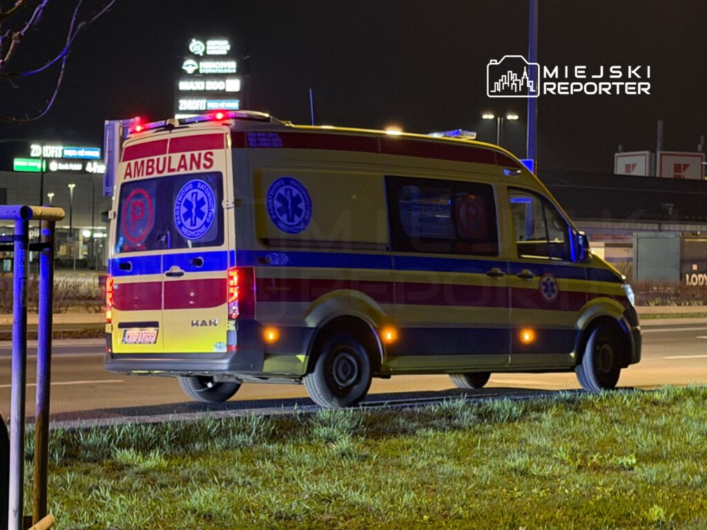Karetka pogotowia z włączonymi światłami awaryjnymi stoi na poboczu drogi nocą, w tle widoczne oświetlenie miejskie.