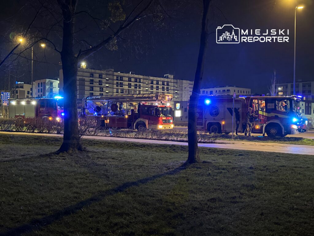 Dwa wozy strażackie z włączonymi sygnałami świetlnymi stoją na ulicy w nocy, obok nowoczesnych budynków mieszkalnych.