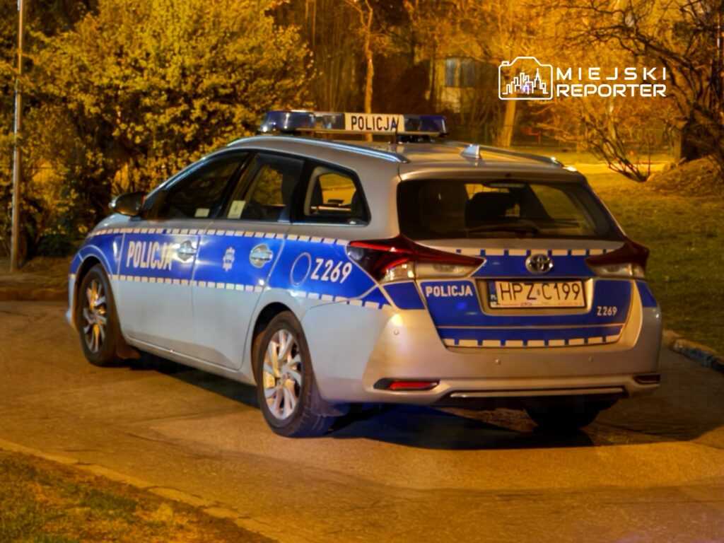 Policyjny samochód osobowy zaparkowany na oświetlonej ulicy, otoczony przez drzewa i krzewy.