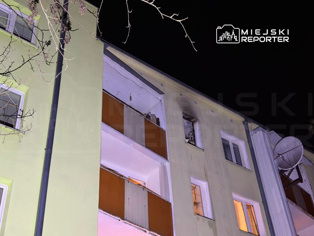 Zniszczona elewacja bloku mieszkalnego z otwartymi oknami i widocznymi uszkodzeniami na wyższych piętrach, nocą.