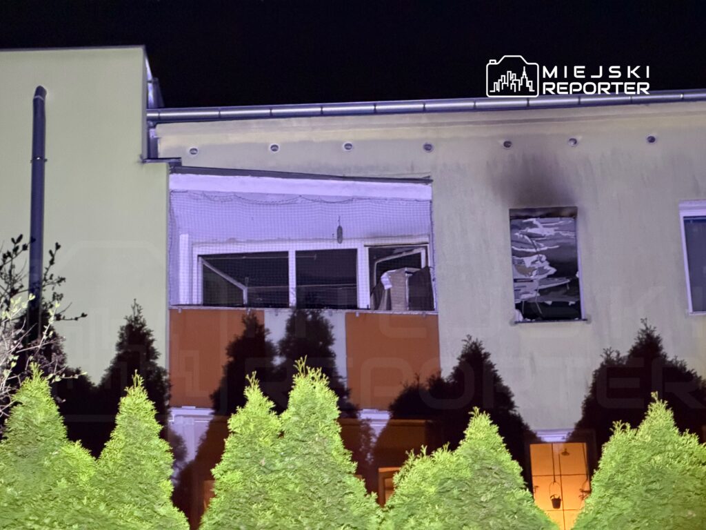 Zniszczony balkon budynku z wybitymi oknami, w tle widoczne iglaki i nocne oświetlenie.