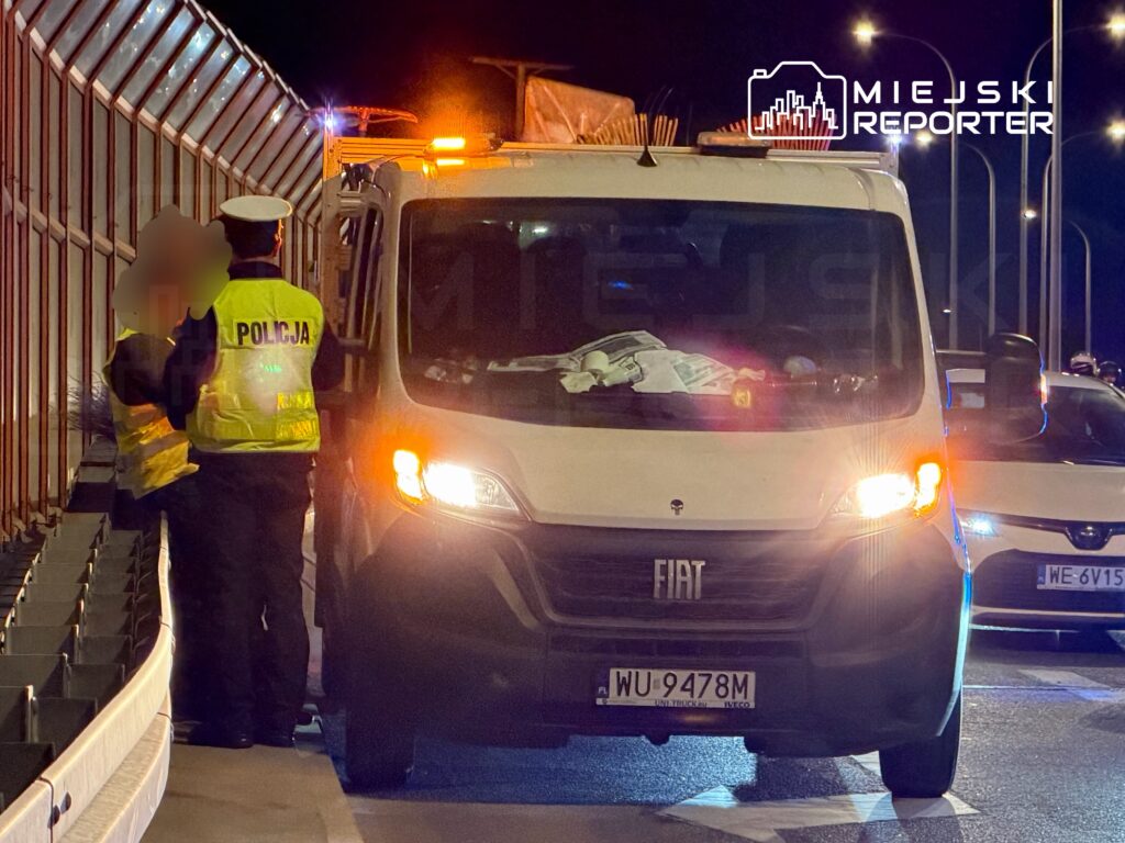 Policjant w kamizelce odblaskowej rozmawia z osobą obok białego furgonu na drodze nocą.