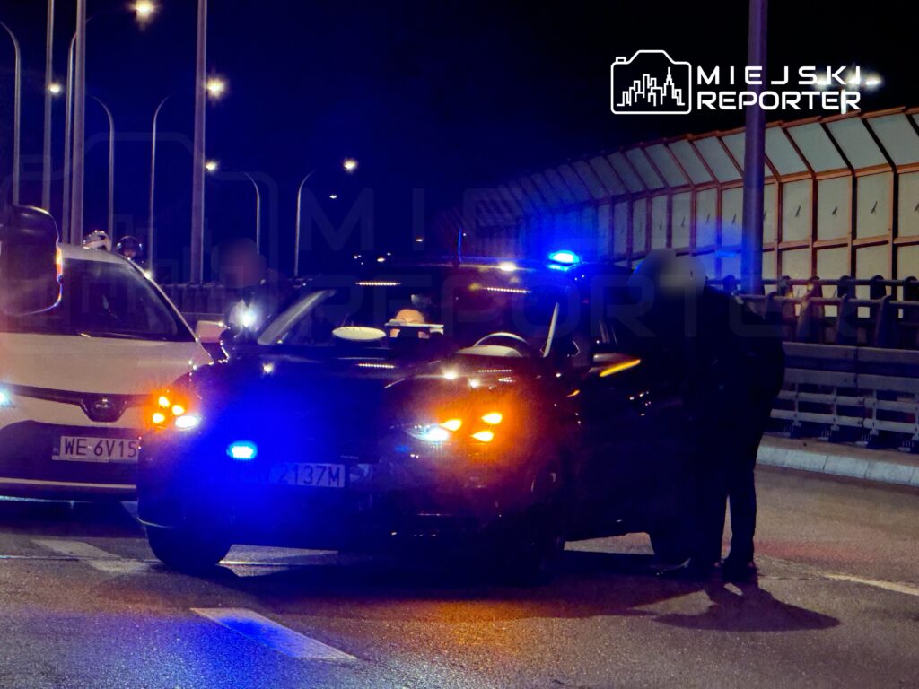 Czarny radiowóz policyjny z włączonymi sygnałami świetlnymi zatrzymuje białe auto na oświetlonej drodze nocą.