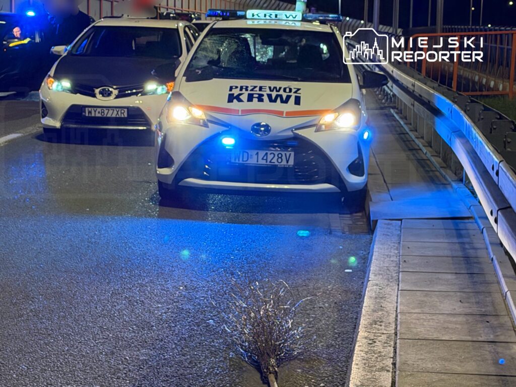 Samochód do przewozu krwi z włączonymi sygnałami świetlnymi stoi na drodze, obok inny pojazd osobowy.