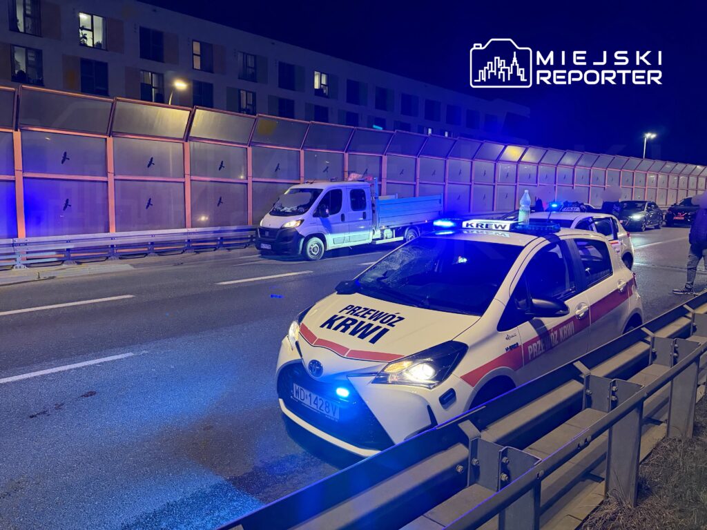 Na jezdni zatrzymane są pojazdy ratunkowe, a w tle widoczny jest nowoczesny budynek z oświetlonymi oknami.