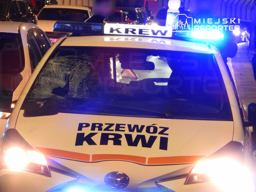 Policyjny samochód z uszkodzoną szybą, zaparkowany w nocy, z włączonymi sygnalizatorami świetlnymi.