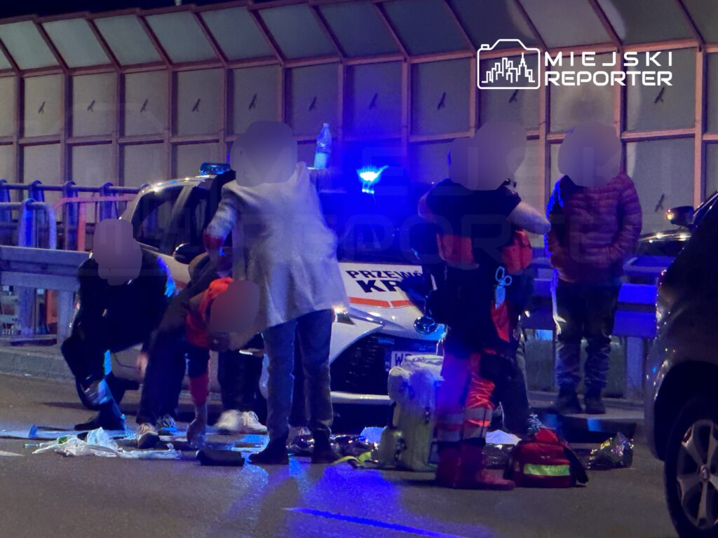 Grupa ratowników medycznych udziela pomocy poszkodowanemu na jezdni, obok radiowozu policyjnego z włączonymi sygnałami świetlnymi.