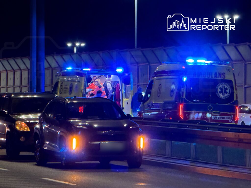 Zespół ratowników medycznych wchodzi do karetki na oświetlonej drodze nocą, otoczonej pojazdami osobowymi.