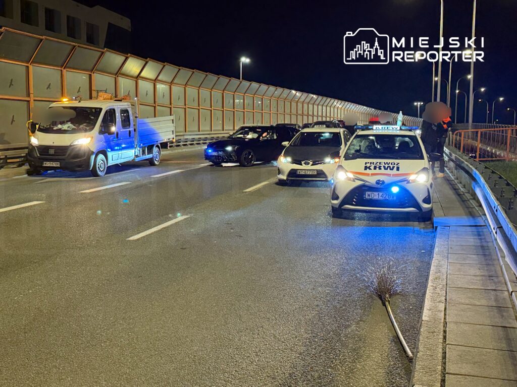 Na drodze nocą zatrzymane są pojazdy, w tym samochód policyjny oraz samochód dostawczy, obok leży miotła.