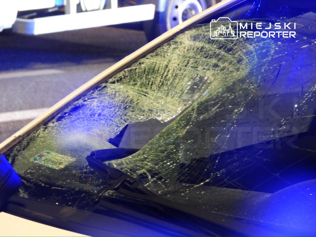 Zniszczona szyba samochodowa z pęknięciami, w tle niebieskie światła policyjne na ulicy.