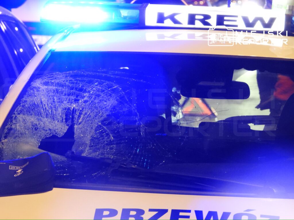 Zniszczona szyba w radiowozie policyjnym oświetlonym niebieskimi światłami.