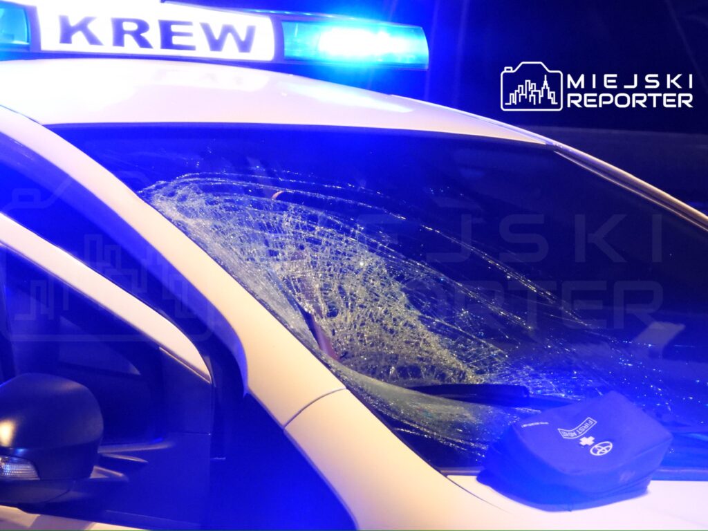 Policyjny radiowóz z uszkodzoną szybą przednią, oświetlony niebieskimi światłami sygnalizacyjnymi.
