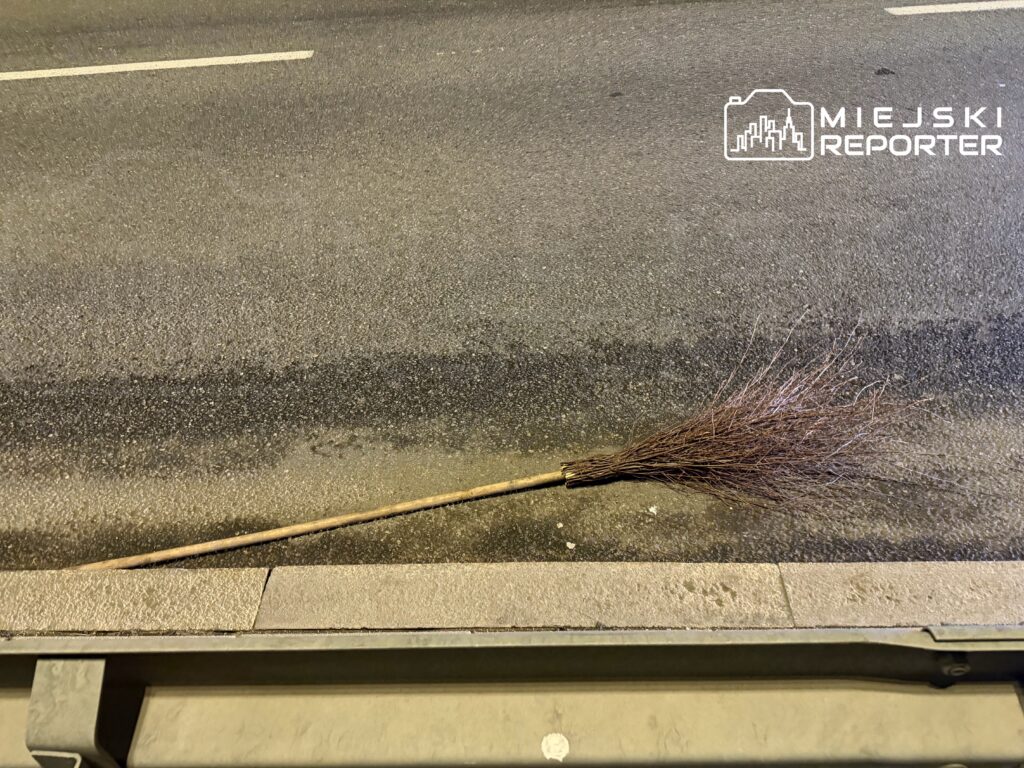 Miotła leżąca na jezdni, obok krawężnika, na asfaltowej drodze.